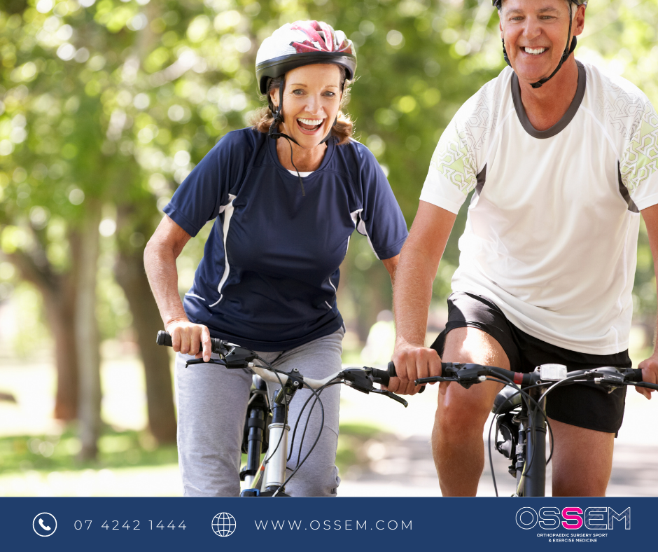 Smiling middle aged male and female riding bicycles surrounded by trees in a park.
